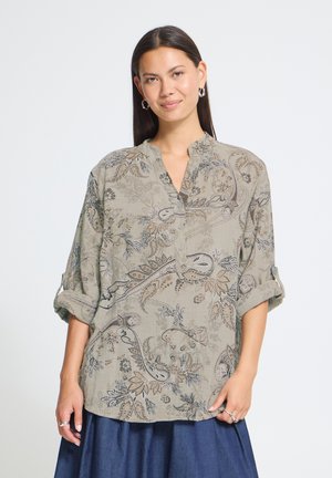 Kvinde med langt mørkt hår, iført sølv hoop-øreringe og en beige paisley-mønstret bluse med op-rullede ærmer og et blåt nederdel.