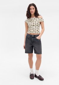 Benäget topp i krämfärg med svarta prickar, kombinerat med mörkgrå denimshorts. Modellen har på sig bruna loafers med vita strumpor.