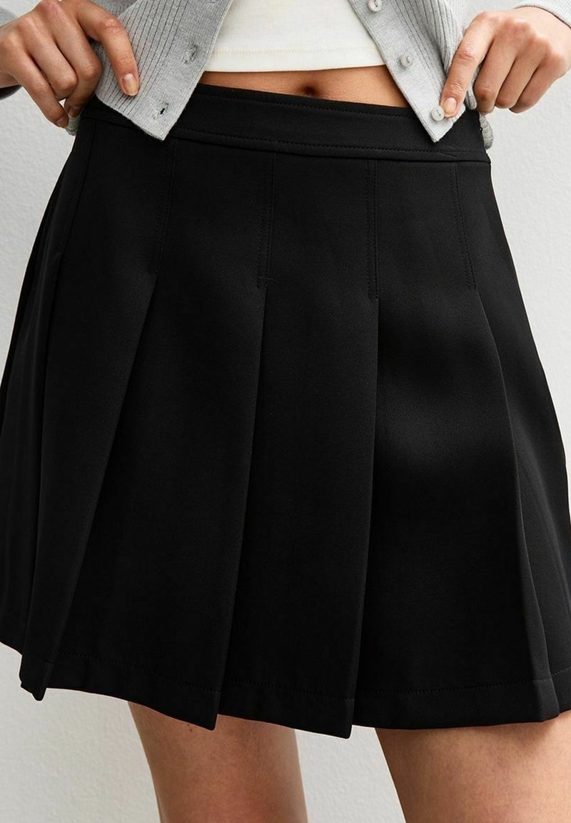 Femme portant une jupe noire plissée taille haute et un gilet gris court avec un haut blanc en dessous.