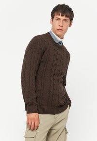Giovane uomo che indossa un maglione a treccia marrone sopra una camicia blu e un dolcevita bianco, con pantaloni cargo beige, in piedi contro uno sfondo bianco.