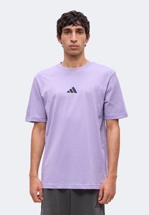 Férfi világoslila Adidas pólóban, melyen fekete logó van a mellkas közepén, semleges arckifejezéssel áll, egyszerű háttér előtt.