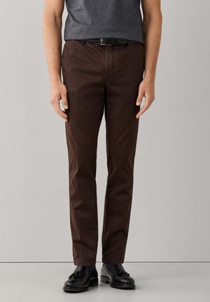 Man die donkere bruine rechte broek draagt met een zwarte riem en zwarte loafers met franjes, staand tegen een effen lichtgrijze achtergrond.