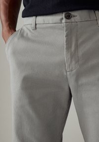 Pantalons ajustés gris clair en tissu légèrement texturé. Comprend des poches latérales, une fermeture à bouton et des passants de ceinture.