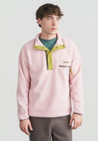 Νεαρός άνδρας που φοράει ροζ φλις πουλόβερ με κίτρινα κουμπιά κλικ και τσέπη με φερμουάρ στο στήθος, στέκεται μπροστά σε απλό φωτεινό φόντο.