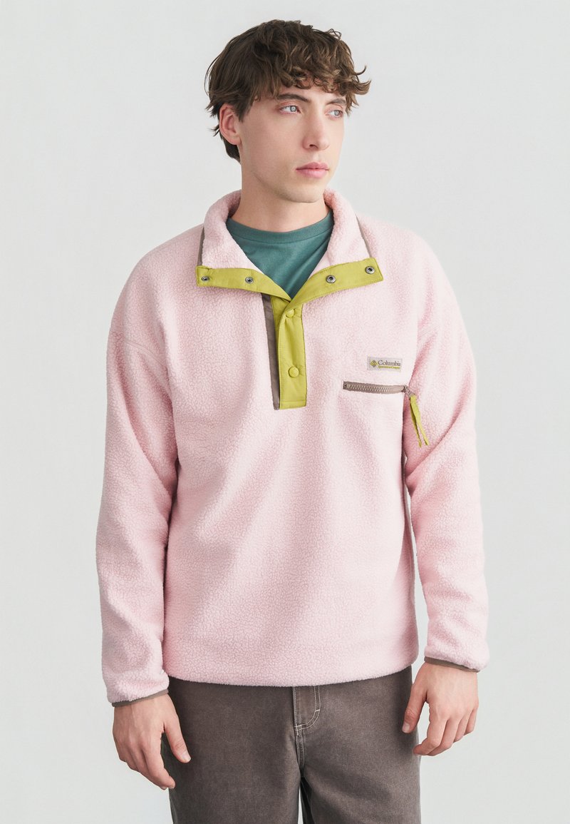 Νεαρός άνδρας που φοράει ροζ φλις πουλόβερ με κίτρινα κουμπιά κλικ και τσέπη με φερμουάρ στο στήθος, στέκεται μπροστά σε απλό φωτεινό φόντο.