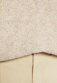 Cremfarbener, strukturierter Strickpullover mit einem kurzen Saum über hellbeigen Hosen, die aus einem glatten Stoff bestehen und sichtbare Nähte aufweisen.