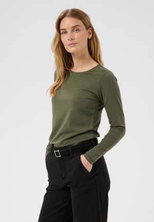 Jeune femme aux longs cheveux blonds portant une chemise à manches longues vert olive et un pantalon noir, mains dans les poches, sur un fond uni.