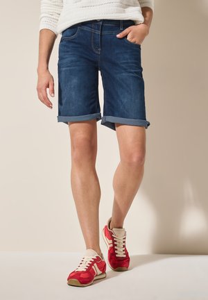 Shorts en denim bleu foncé avec ourlets roulés, associés à des chaussures de sport rouges avec des accents blancs et des détails beiges. Haut en tricot simple.