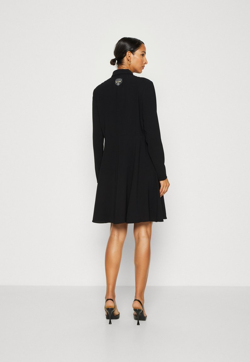 Schwarzes langärmliges Kleid mit tailliertem Oberteil und ausgestelltem Rock, ausgestattet mit einem Reißverschluss hinten und Logodetail. Getragen mit schwarzen Riemchensandalen.