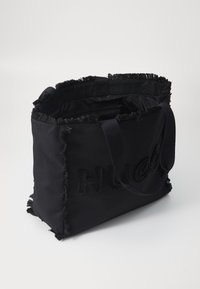 Crna platnena torba s razderanim rubovima, s uzdignutim logotipom "HUGO". Ima čvrste naramenice i prostrano, neobloženo unutrašnjost.