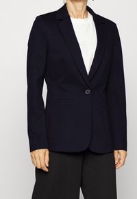 Blazer navy con cierre de un solo botón, solapas con muesca y dos bolsillos frontales, combinado con una camiseta blanca y pantalones negros.
