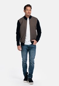 Heren bomberjack met een bruine romp, marineblauwe mouwen, ribgebreide manchetten en kraag. Gepaard met een wit T-shirt en blauwe jeans, afgewerkt met zwarte sneakers.