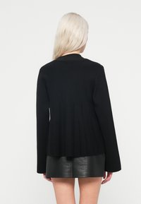 Cardigan negru cu spate evazat, textură canalată și mâneci lungi. Asortat cu o fustă mini neagră cu model de crocodil.