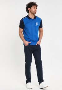Blå pikétröja med marinblå ärmar, prydd med en vit logotyp och tunna vita ränder. Kombinerad med mörka byxor och vita sneakers.