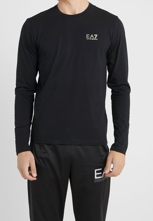 Mann trägt ein schwarzes Langarmshirt und eine schwarze Hose mit kleinen weißen EA7 Emporio Armani Logos auf der Brust und dem Oberschenkel.