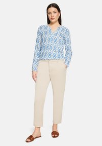 Blouse bleu clair à motifs avec un col en V, associée à un pantalon beige ajusté et des sandales marron à enfiler. La blouse présente un motif floral.