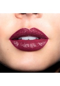 Rouge à lèvres prune foncé avec une finition brillante, appliqué sur des lèvres pleines, mettant en valeur une texture lisse et une forme définie contre des dents blanches.