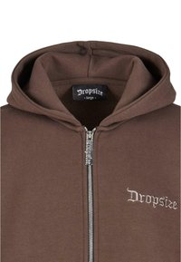 Smeđa zip-up dukserica izrađena od mješavine pamuka, sa prednjim zatvaračem i izvezenim logom "Dropsize" na lijevoj strani grudi.
