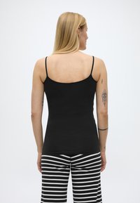 Ženska s svetlimi lasmi, oblečena v črno majico s tankimi naramnicami in črno-bele črtaste hlače, ki kaže tetovaže na desni roki.