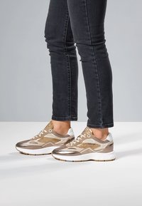 Sportliche Sneaker in metallischem Gold und Weiß, mit Mesh, Glitzerdetails und einer klobigen weißen Sohle. Kombiniert mit eng anliegenden schwarzen Jeans.