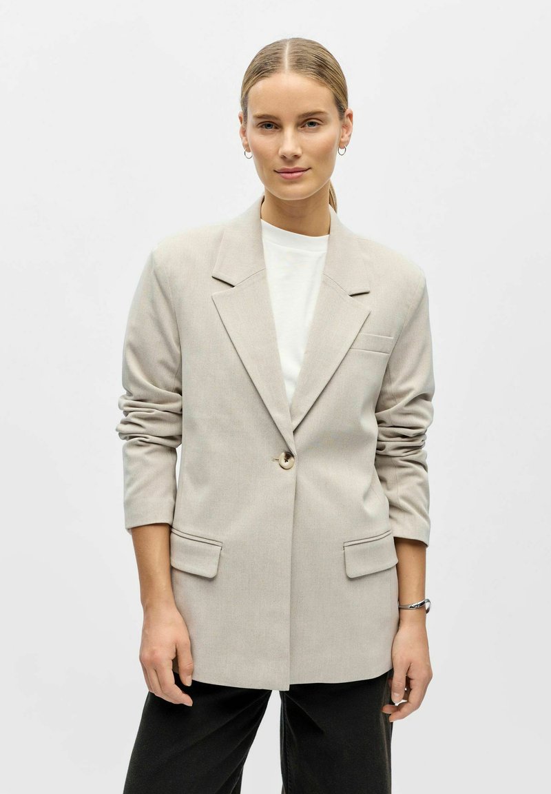 Mujer joven con el cabello peinado hacia atrás lleva un blazer beige de un solo botón sobre una camisa blanca y pantalones negros, de pie frente a un fondo blanco liso.