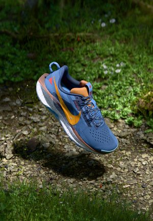 Blue mesh sneaker with orange accents and a yellow logo, featuring a speckled white sole and textured lace area, floating above a natural backdrop.