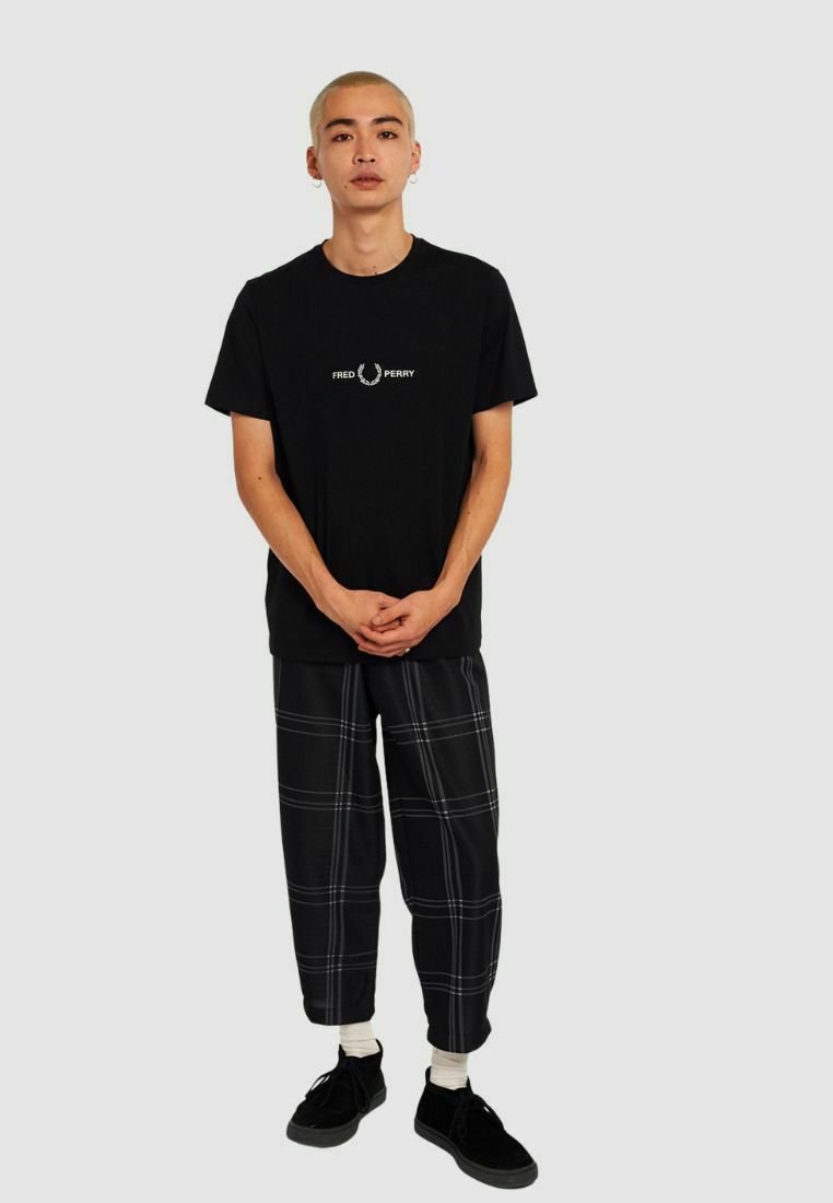 Camisa de algodón negra con el logo blanco de Fred Perry, combinada con pantalones cuadros negros que presentan líneas blancas. El modelo lleva zapatos negros y calcetines blancos.