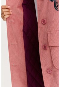 Ružový kordúrový kabát s prešívanou fialovou podšívkou, obsahujúci štyri gombíky, vrecko a detailné prešívanie na vonkajšej strane.