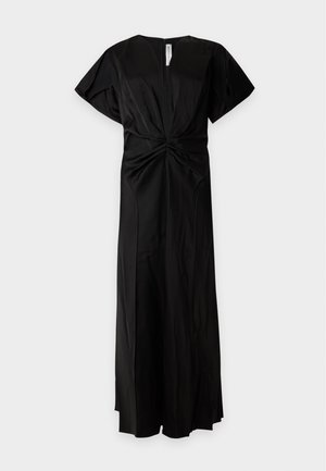 Femme aux longs cheveux foncés portant une robe noire à col en V avec des manches courtes, une taille froncée et des sandales noires à talons hauts sur fond blanc.