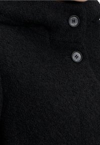 Cappotto nero con texture e colletto alto, caratterizzato da due bottoni neri con la scritta "Gil Bret" in rilievo. Il tessuto appare caldo e accogliente.