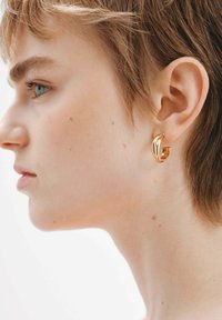 Pendiente de aro de metal dorado con un diseño retorcido. Mostrado en un cabello corto con un tono de piel claro y textura de piel visible.