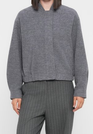 Personne portant une veste courte gris moyen à manches longues et un pantalon gris foncé à fines rayures.