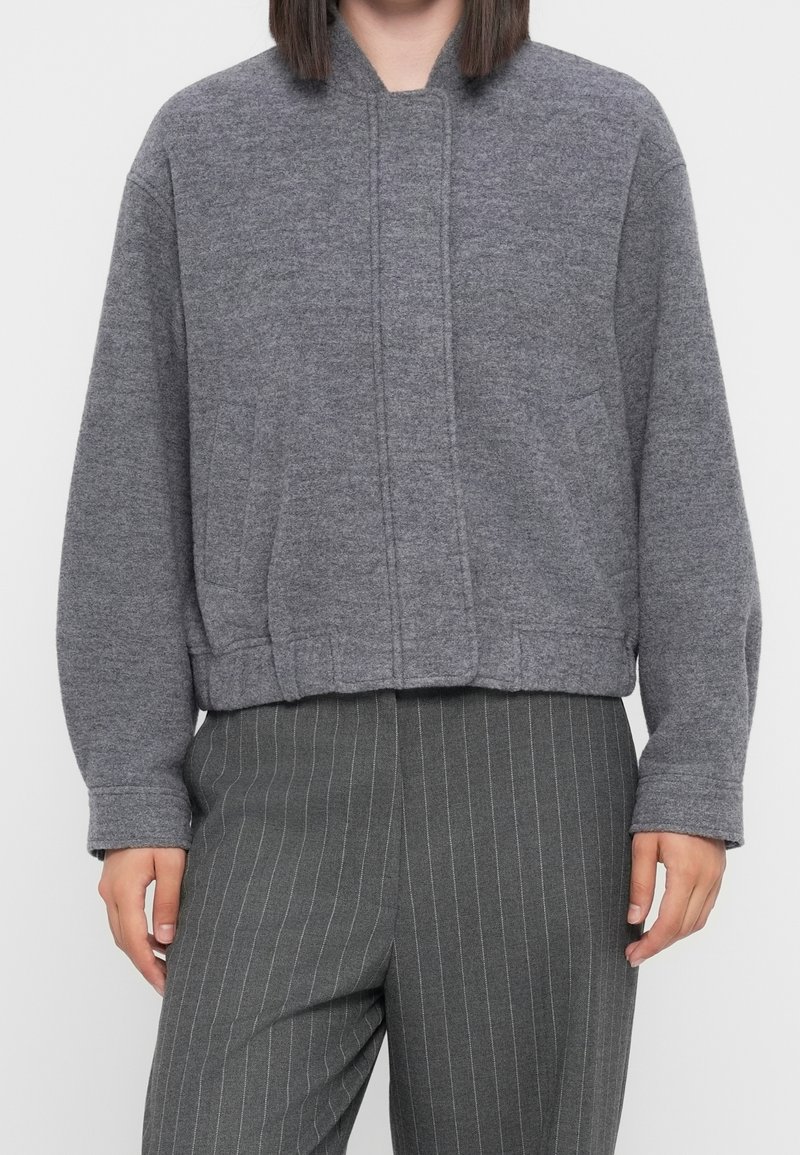 Personne portant une veste courte gris moyen à manches longues et un pantalon gris foncé à fines rayures.