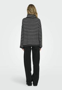 Frau, die sich mit dem Rücken zur Kamera dreht, trägt einen grau-schwarz gestreiften Rollkragenpullover und lockere schwarze Hosen vor einem schlichten hellen Hintergrund.