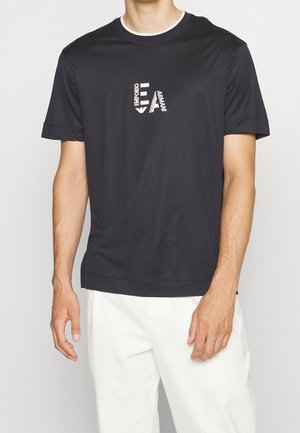 Mężczyzna w czarnej koszulce Emporio Armani z białym logo na piersi i białych spodniach, stojący na jednolitym tle.