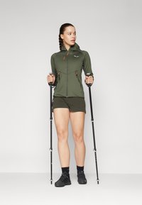 Olivengrønn zip-up jakke med oransje detaljer, kombinert med mørke shorts. Brukeren holder turstaver, iført svarte tursko og sokker.
