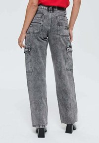 Personne portant un jean cargo gris délavé taille haute avec une ceinture noire et des bottes à talons noires, se tenant devant un fond clair.