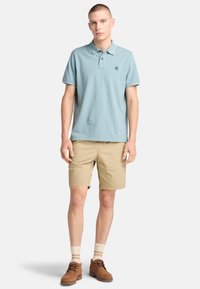 Ljusblå pikétröja med korta ärmar, tillsammans med beige shorts. Bruna mockaskor, strumpor med krämfärgade ränder och en liten logo på bröstet.