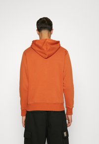 Felpa con cappuccio arancione, realizzata in misto cotone. Presenta una vestibilità ampia, cuciture visibili e polsini a coste. Indossata con pantaloni cargo scuri.