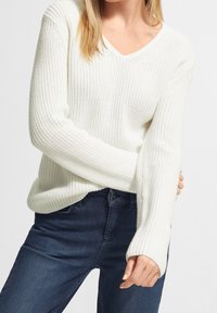 Biały sweter dzianinowy z dekoltem w serek i długimi rękawami, z teksturowanym wzorem i ściągaczami przy mankietach, w zestawie z ciemnoniebieskimi dżinsami.