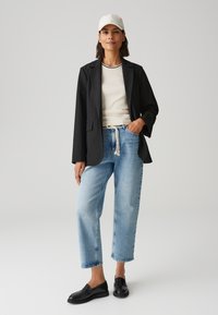 Zwarte blazer over een crèmekleurig topje, lichtblauwe brede jeans en zwarte loafers. Het model draagt een witte pet, met zichtbare zakken op de jeans.