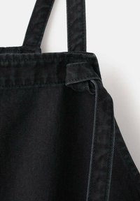 Die schwarze Jeans-Schürze besteht aus robustem Stoff, hat verstellbare Träger und ein gebundenes Knotendetail. Die Oberfläche zeigt eine strukturierte Optik mit leichtem Glanz.