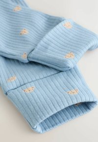 Tela de punto acanalado azul claro con pequeñas caras de ositos beige impresas uniformemente en la superficie, doblada para mostrar textura y patrón.