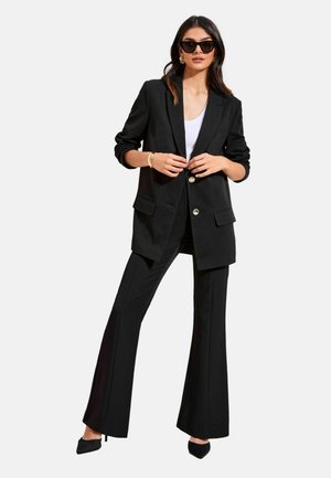 Frau trägt schwarzen übergroßen Blazer mit goldenen Knöpfen, schwarze ausgestellte Hose, weißes Oberteil, schwarze High Heels, goldenes Armband und schwarze Sonnenbrille.