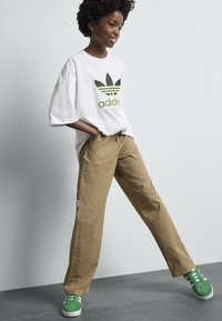 Leende person iklädd en vit oversized Adidas T-shirt, beige vida byxor och gröna sneakers som står mot en enkel ljus vägg.