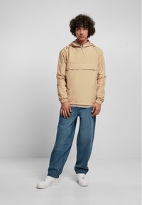 Beige hættepulloverjakke med en lynlås foran, parret med afslappede blå jeans og hvide sneakers. Glat stof og afslappet pasform.
