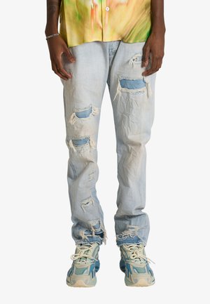 Persona che indossa jeans azzurri strappati con toppe blu e sneakers blu e beige, in piedi con le mani rilassate lungo i fianchi.