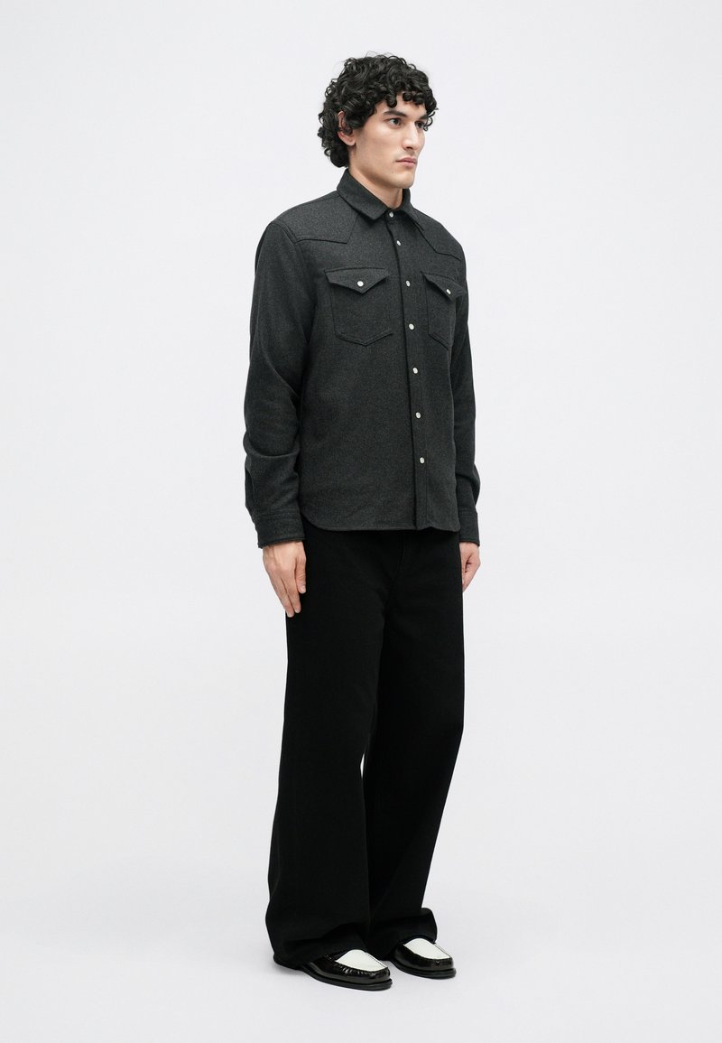 Dark gray wool shirt with two chest pockets, snap buttons, and a structured collar; paired with black wide-leg trousers and black shoes.