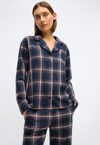 Merellinen ruudullinen pyjamapuku pehmeää puuvillaa, jossa on pitkät hihat, nappikiinnitys ja kontrastina punaisia ja valkoisia viivoja. Pelkistetty kaulus ja tasku täydentävät kokonaisuutta.