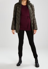 Ocieplana oliwkowa kurtka o pikowanej strukturze, zestawiona z bordową bluzką związaną w talii, czarnymi rurkami oraz botkami na kostkę.
